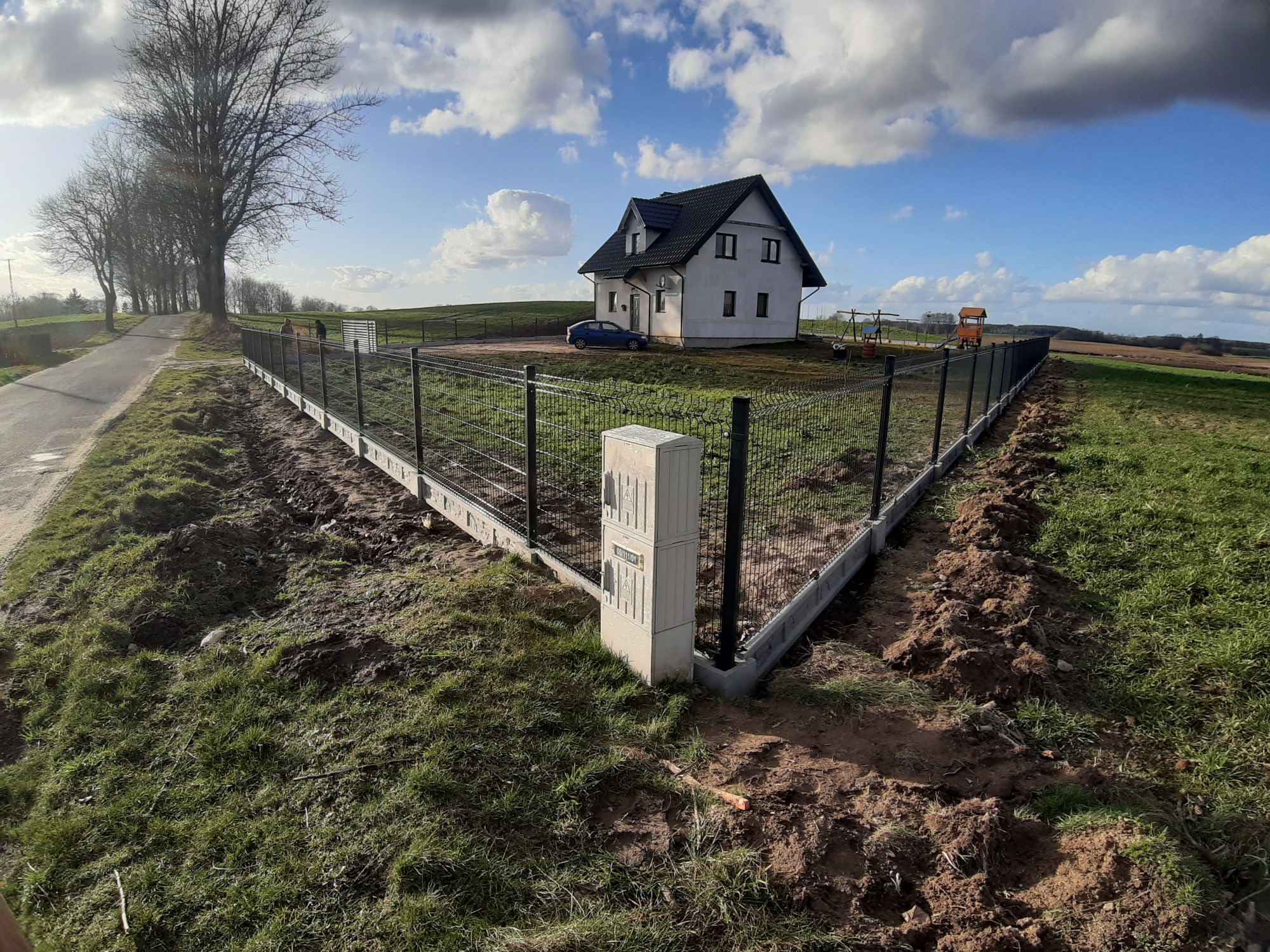 Nowoczesne ogrodzenie panelowe z podmurówką prefabrykowaną, otaczające posesję z domem jednorodzinnym i placem zabaw w tle, widok z perspektywy ulicy w słoneczny dzień.