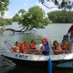 Grupa dzieci w pomarańczowych kamizelkach ratunkowych płynie łodzią motorową po jeziorze w słoneczny dzień, pod nadzorem dorosłych.