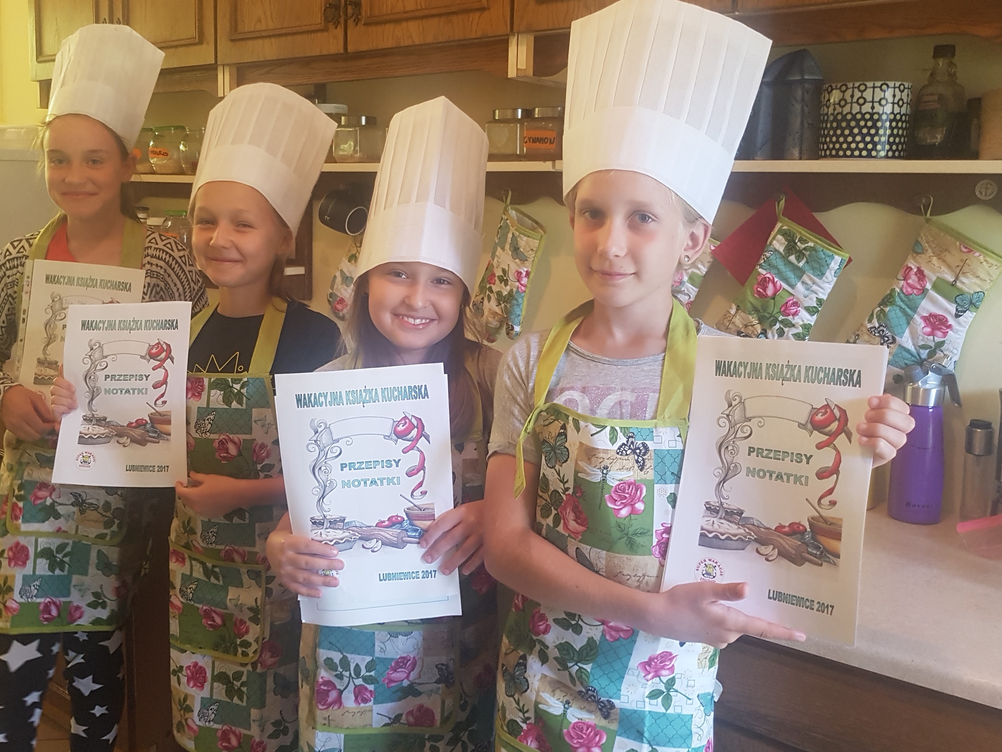 Grupa uśmiechniętych dzieci w kucharskich czapkach i fartuchach trzymających książki kucharskie z napisem 'Wakacyjna Książka Kucharska - Przepisy Notatki Lubniewice 2017'.