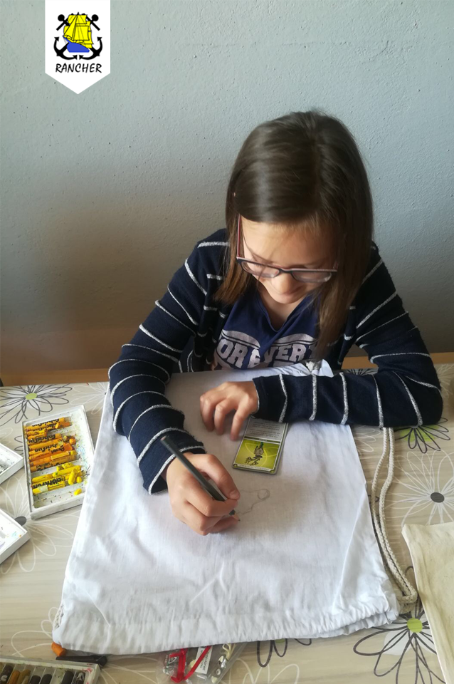 Dziewczynka w okularach rysuje ołówkiem na białym worku, używając karty jako wzoru, obok leżą kredki pastelowe.