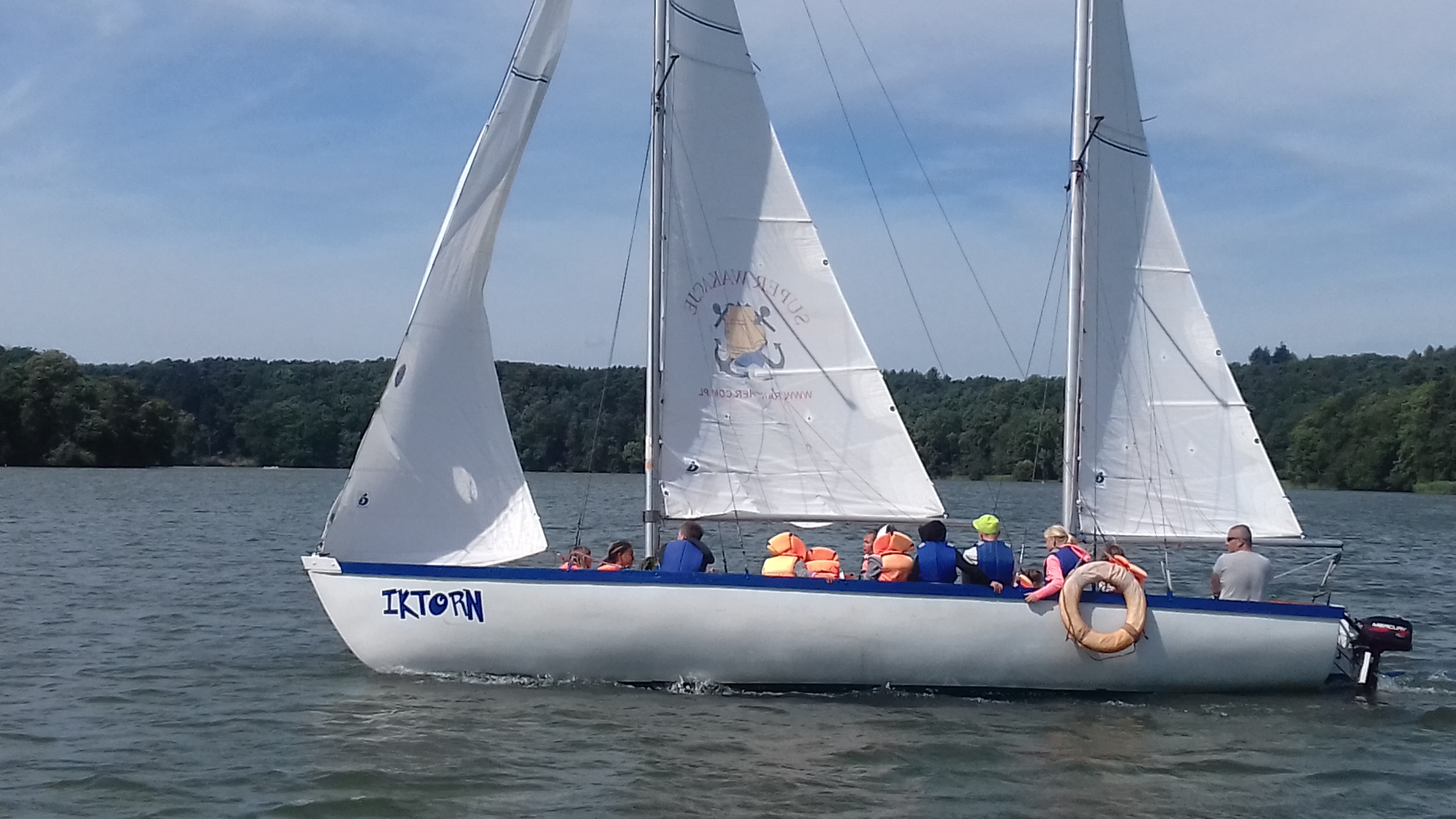 Żaglówka IKTORN z grupą dzieci w kamizelkach ratunkowych płynie po jeziorze w słoneczny dzień, widoczne lasy na brzegu.