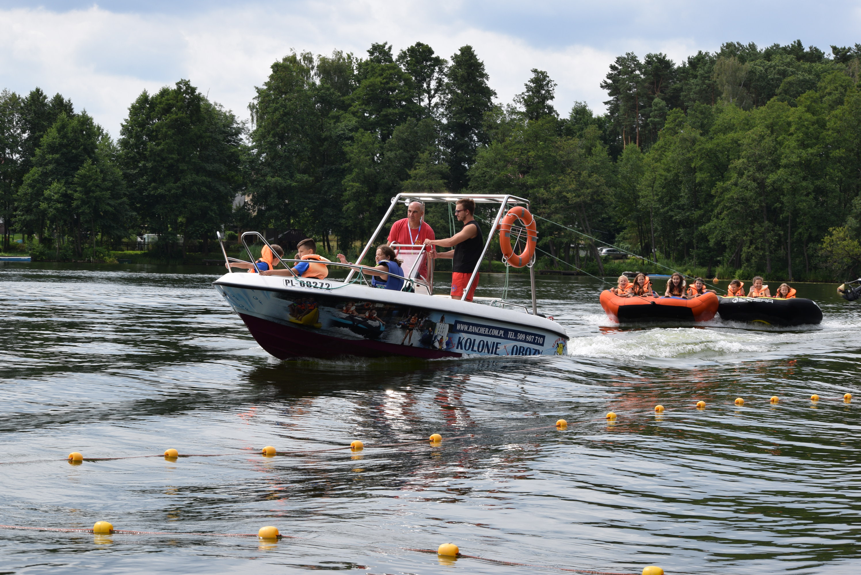 Motorówka ciągnie za sobą dmuchany ponton z grupą dzieci w kamizelkach ratunkowych po jeziorze, w tle drzewa.
