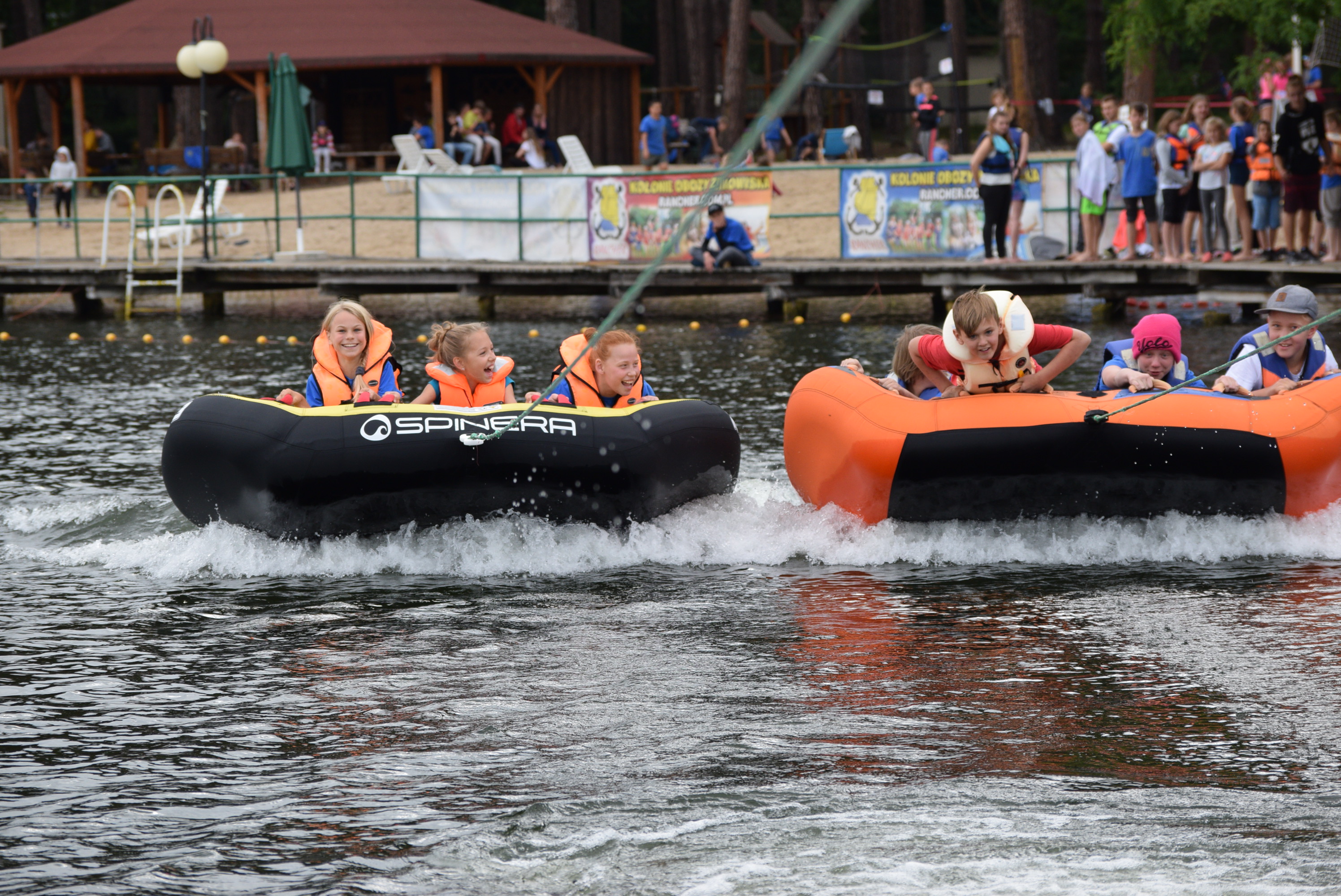 Dwie grupy dzieci w kamizelkach ratunkowych, ciągnięte na dmuchanych pontonach po jeziorze, z uśmiechem na twarzach, na tle pomostu z obserwującymi osobami i banerami reklamowymi.