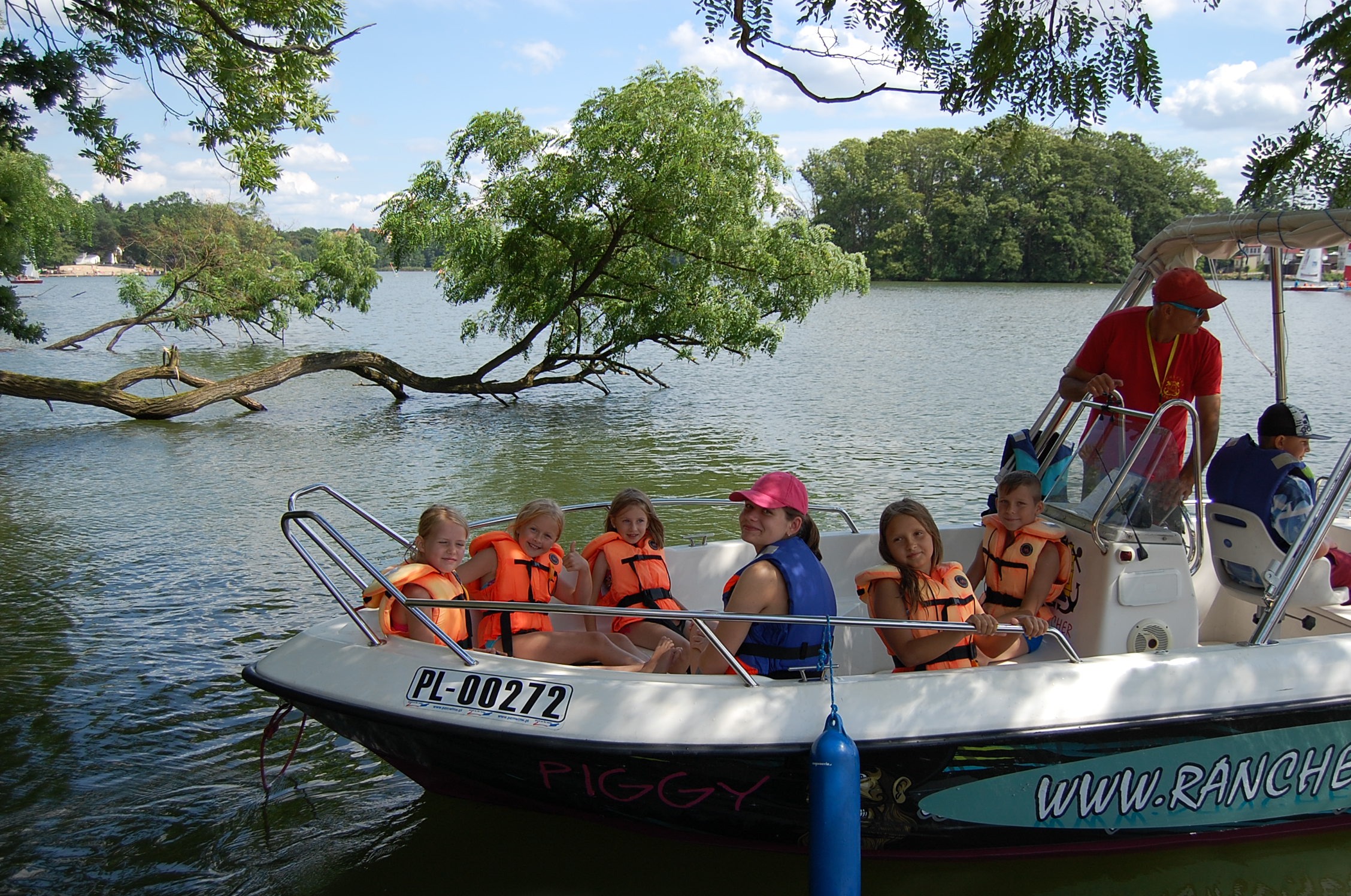 Grupa dzieci w pomarańczowych kamizelkach ratunkowych płynie łodzią motorową po jeziorze w słoneczny dzień, pod nadzorem dorosłych.