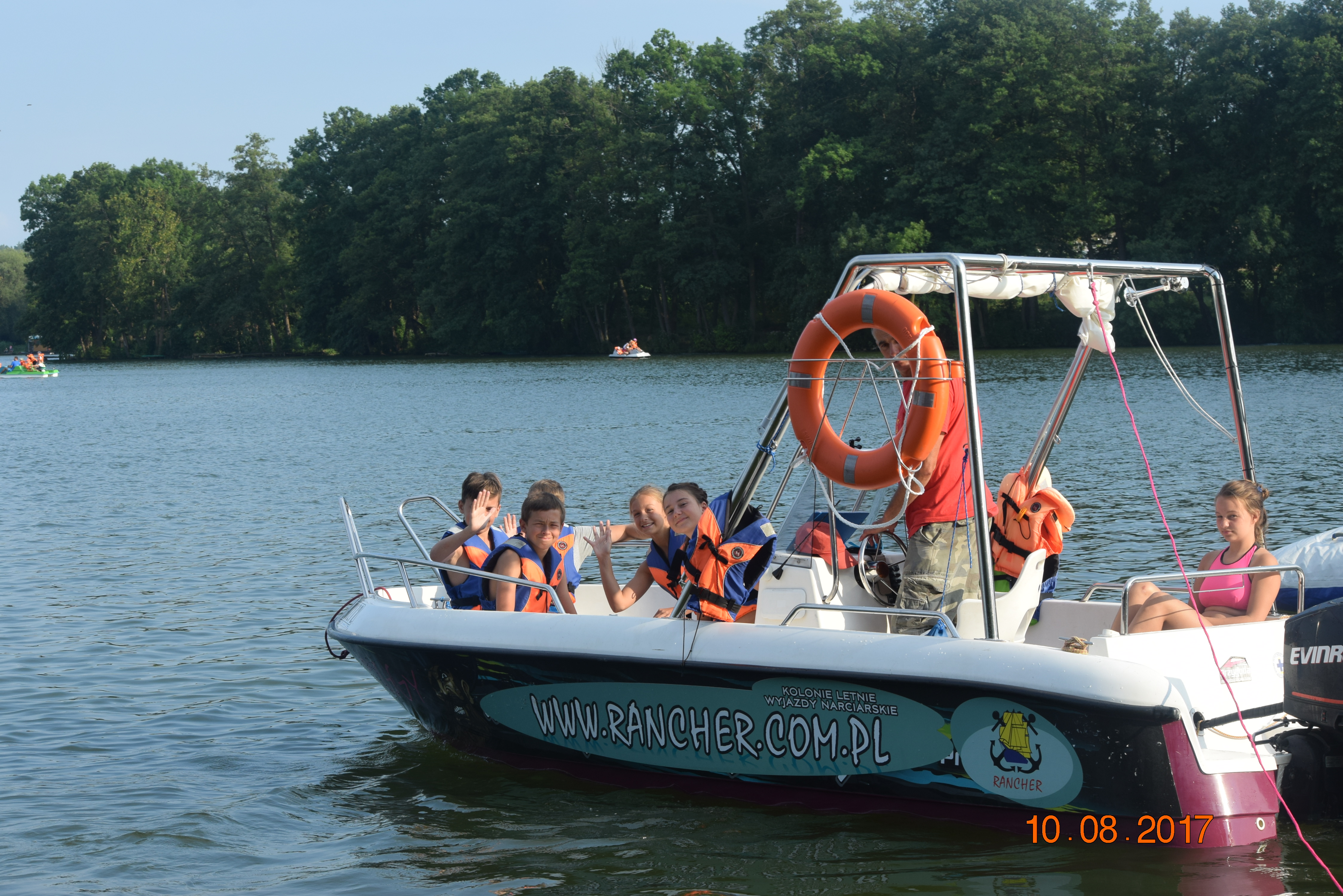 Motorówka z uśmiechniętymi dziećmi w kamizelkach ratunkowych na jeziorze, prowadzona przez sternika, z widocznym kołem ratunkowym i reklamą firmy Rancher.com.pl na burcie.