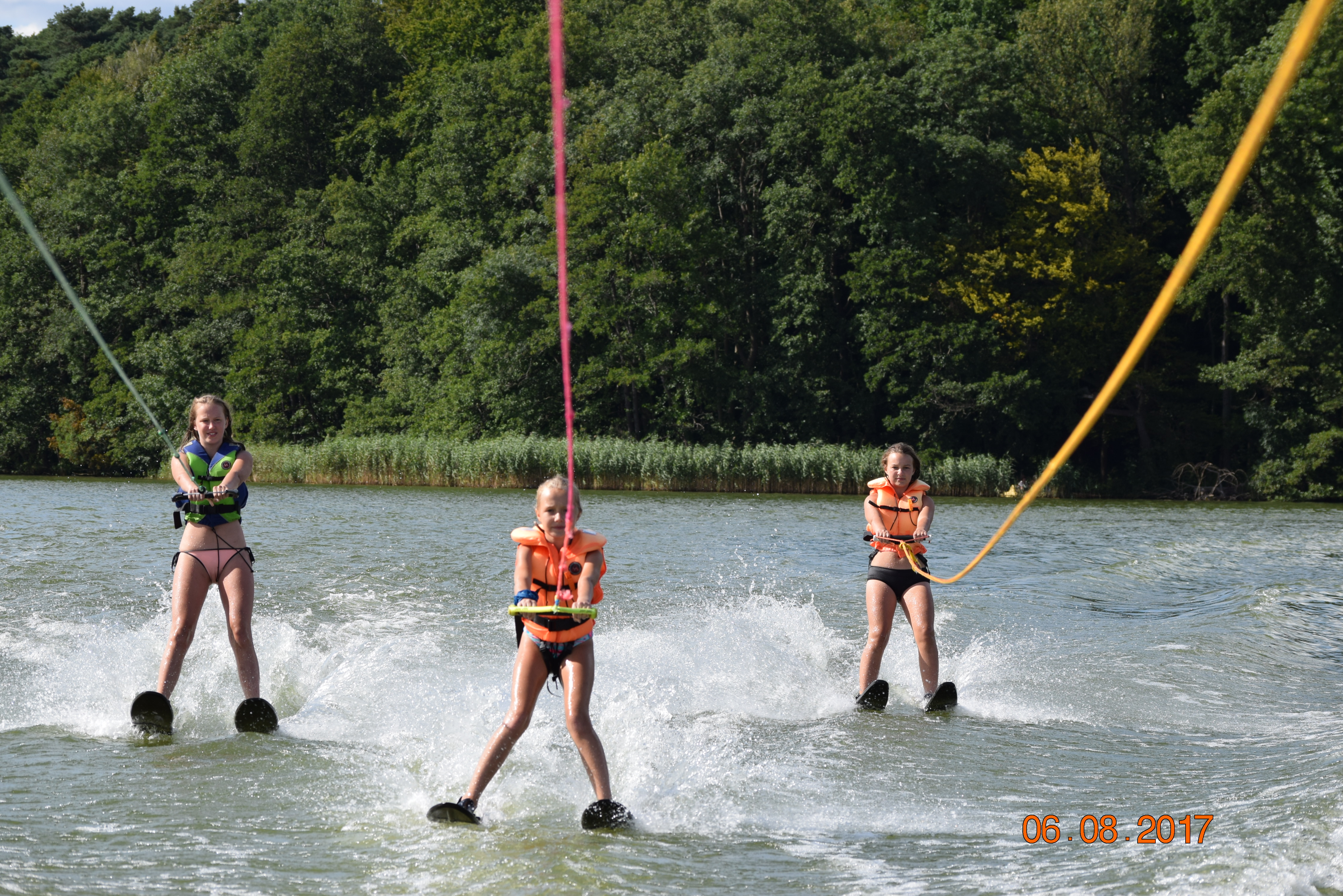 Trzy dziewczyny na nartach wodnych ciągnięte przez motorówkę na tle zielonego lasu, widoczne rozpryski wody i liny holownicze.