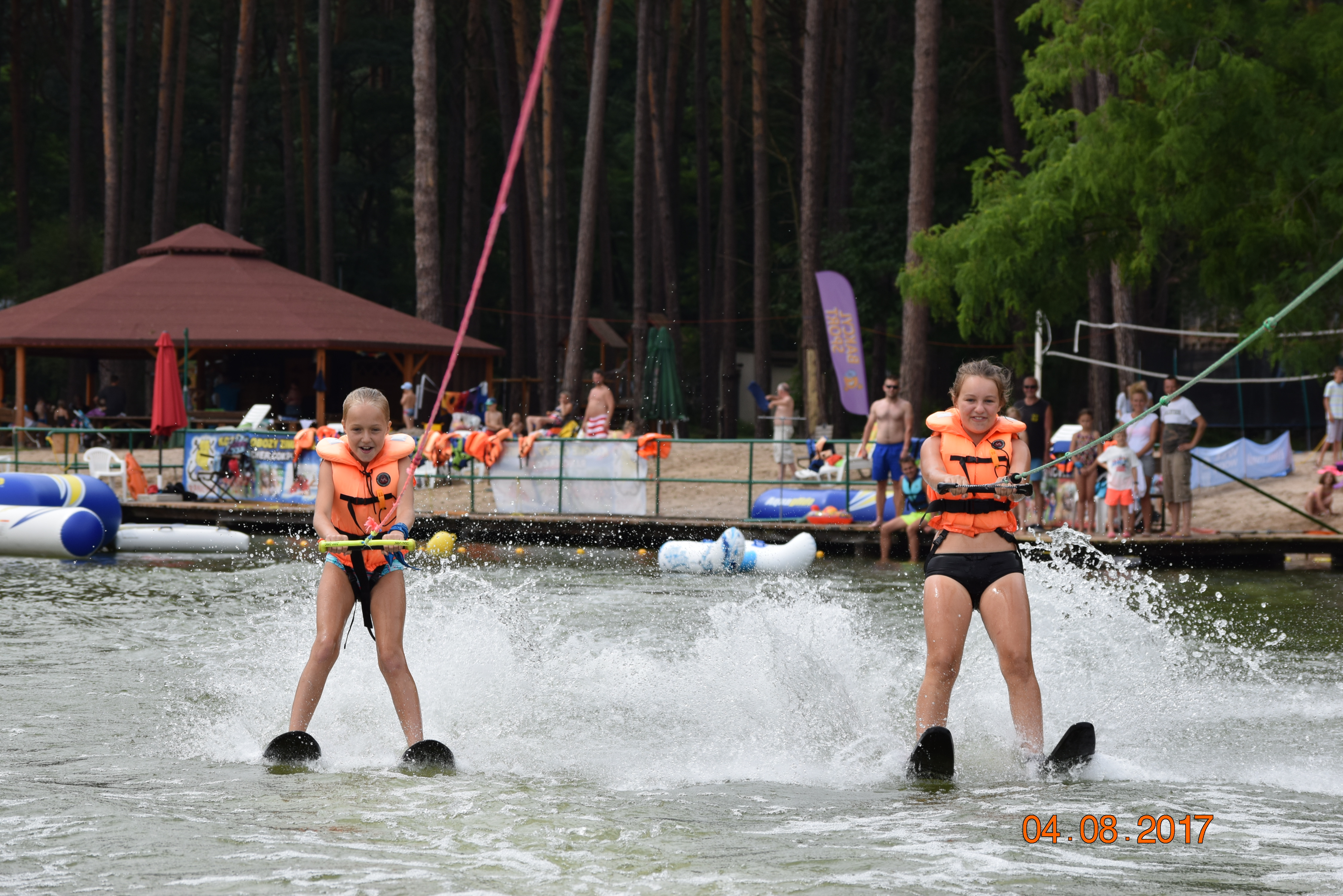 Dwie dziewczynki w pomarańczowych kamizelkach asekuracyjnych na nartach wodnych, ciągnięte liną po jeziorze, w tle plaża z ludźmi i las.