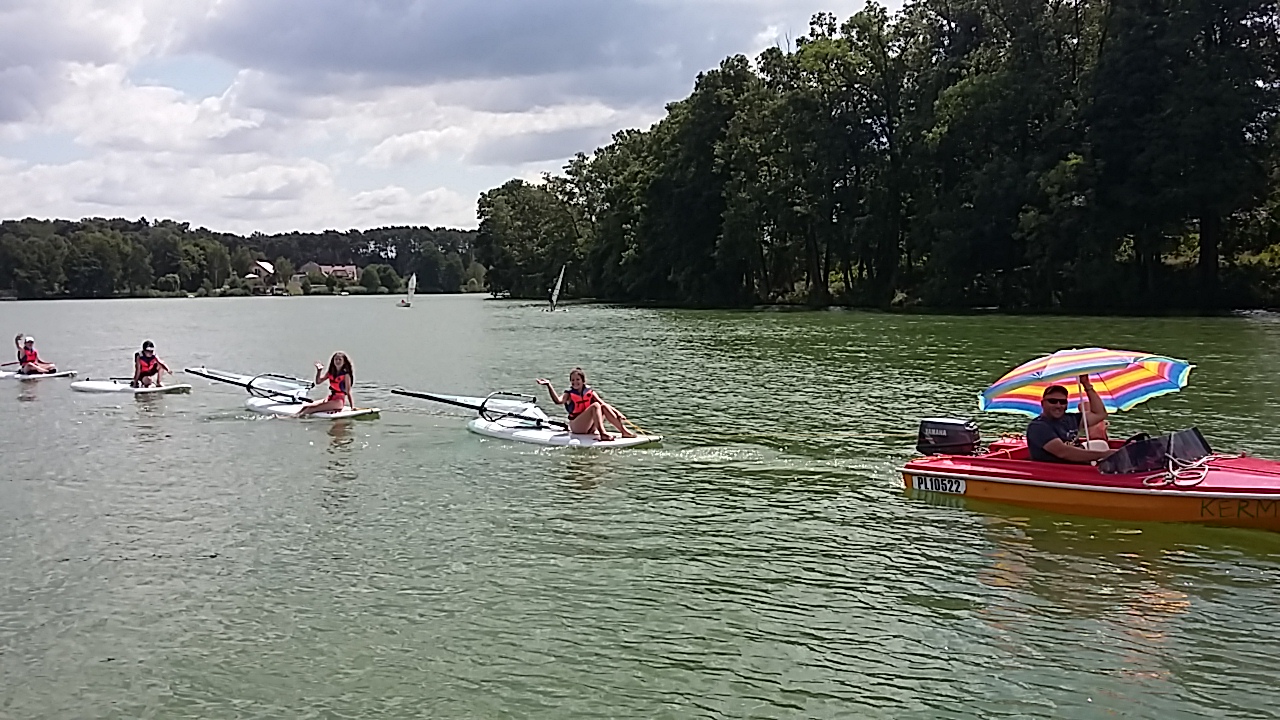 Grupa dzieci na deskach windsurfingowych holowanych przez łódź motorową z kolorowym parasolem na jeziorze, w tle zielony las i budynki.