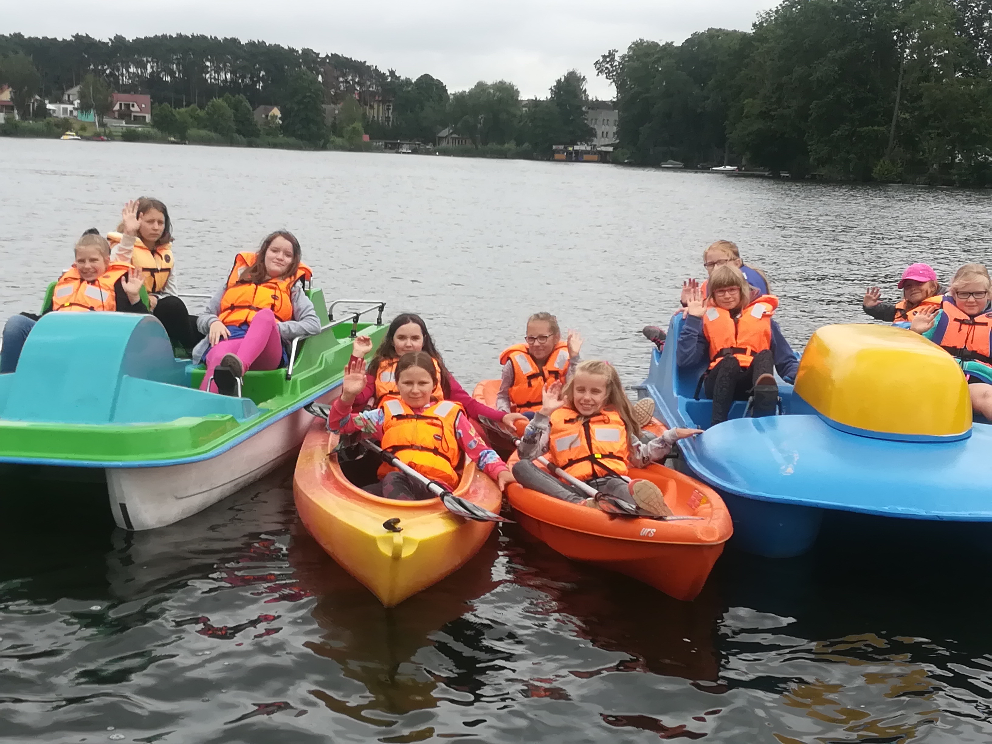 Grupa dzieci w kamizelkach ratunkowych macha z kolorowych kajaków i rowerków wodnych na jeziorze, w tle widoczny las.