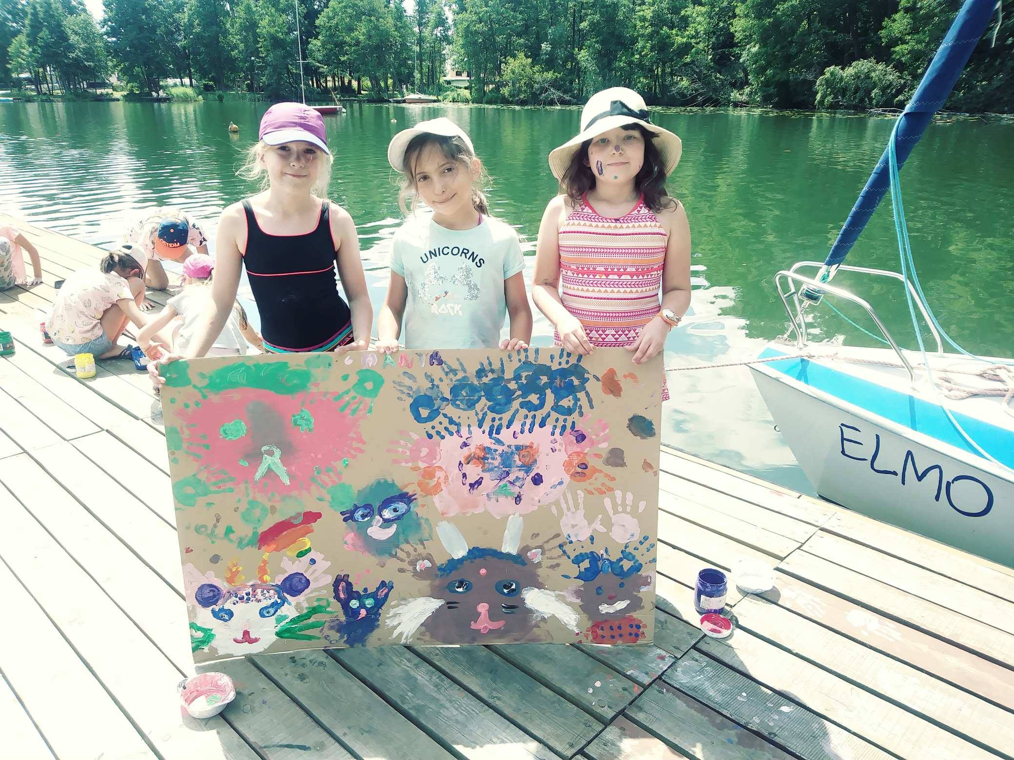 Trzy dziewczynki trzymają karton z malunkami dłoni i zwierząt na drewnianym pomoście nad jeziorem, obok zacumowana łódka z napisem ELMO.