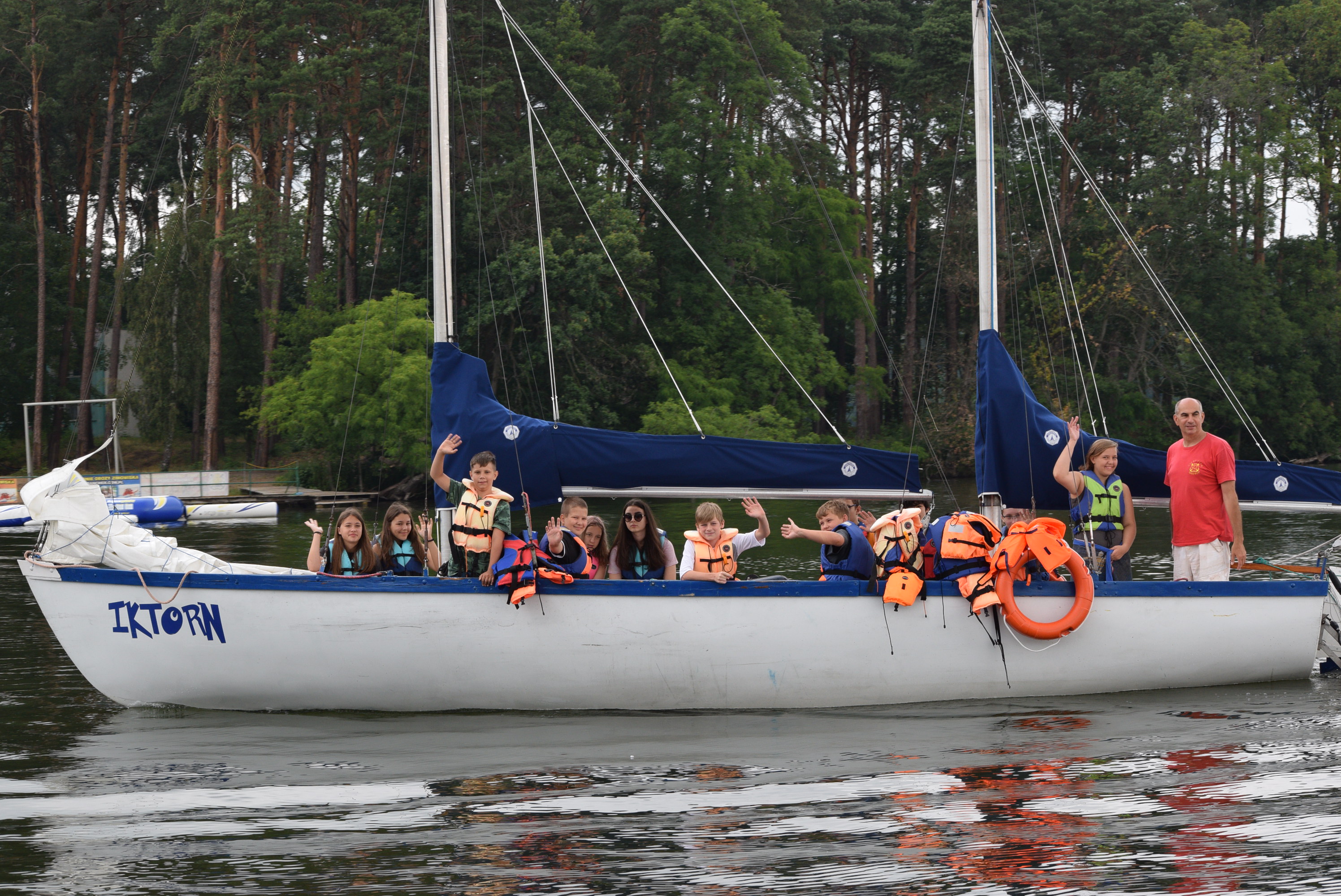 Grupa dzieci w kamizelkach ratunkowych macha z pokładu białej żaglówki 'IKTORN' na tle zielonego lasu i jeziora.