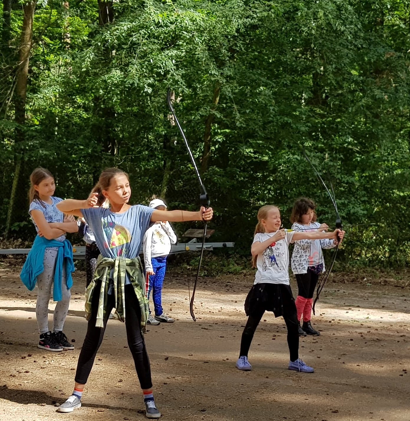 Grupa dziewcząt ćwiczy strzelanie z łuku na leśnej polanie, koncentrując się na celowaniu.