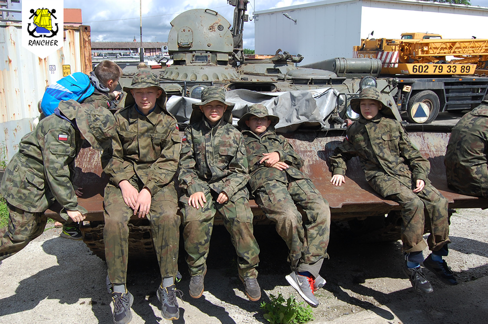 Grupa dzieci w mundurach siedzi na opancerzonym pojeździe wojskowym; w tle kontener, dźwig i budynek.