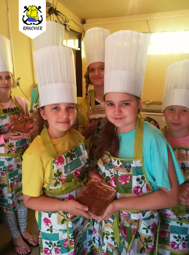 Grupa dzieci w czapkach kucharskich i fartuchach prezentuje upieczone ciasta w pomieszczeniu o żółtych ścianach.