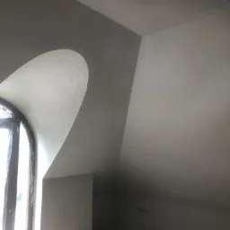 Poddasze po nałożeniu gładzi, widoczny fragment okna z łukowym nadprożem, sufit skośny i ściany przygotowane do malowania.