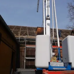 Żuraw wieżowy podnosi materiały na remontowany dach budynku gospodarczego, widoczni dwaj pracownicy na dachu.