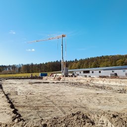 Z.U.B. Sklorz - Budowa w toku: teren przygotowany pod fundamenty, widoczny dźwig, koparka i budynek w tle. Słoneczny dzień, niebieskie niebo.