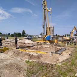 Z.U.B. Sklorz - Budowa fundamentów domu: wykop, szalunki, zbrojenie, beton. Na budowie dźwig, koparka i betoniarka. Widoczni pracownicy. Słoneczny dzień, błękitne niebo.