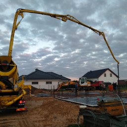 Z.U.B. Sklorz - Żółta betonomieszarka z wysięgnikiem pompuje beton do fundamentów domu jednorodzinnego. W tle widoczne domy i koparka na tle pochmurnego nieba.
