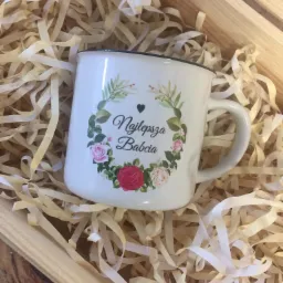 Biały emaliowany kubek z napisem 'Najlepsza Babcia' i motywem kwiatowym, umieszczony w drewnianej skrzynce wypełnionej dekoracyjnym papierem w formie wstążek.