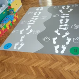 Szary dywan z białymi nadrukami stóp i dłoni, oznaczony numerami 1 i 2, leżący na parkiecie w sali przedszkolnej z kolorowymi meblami i dekoracjami.