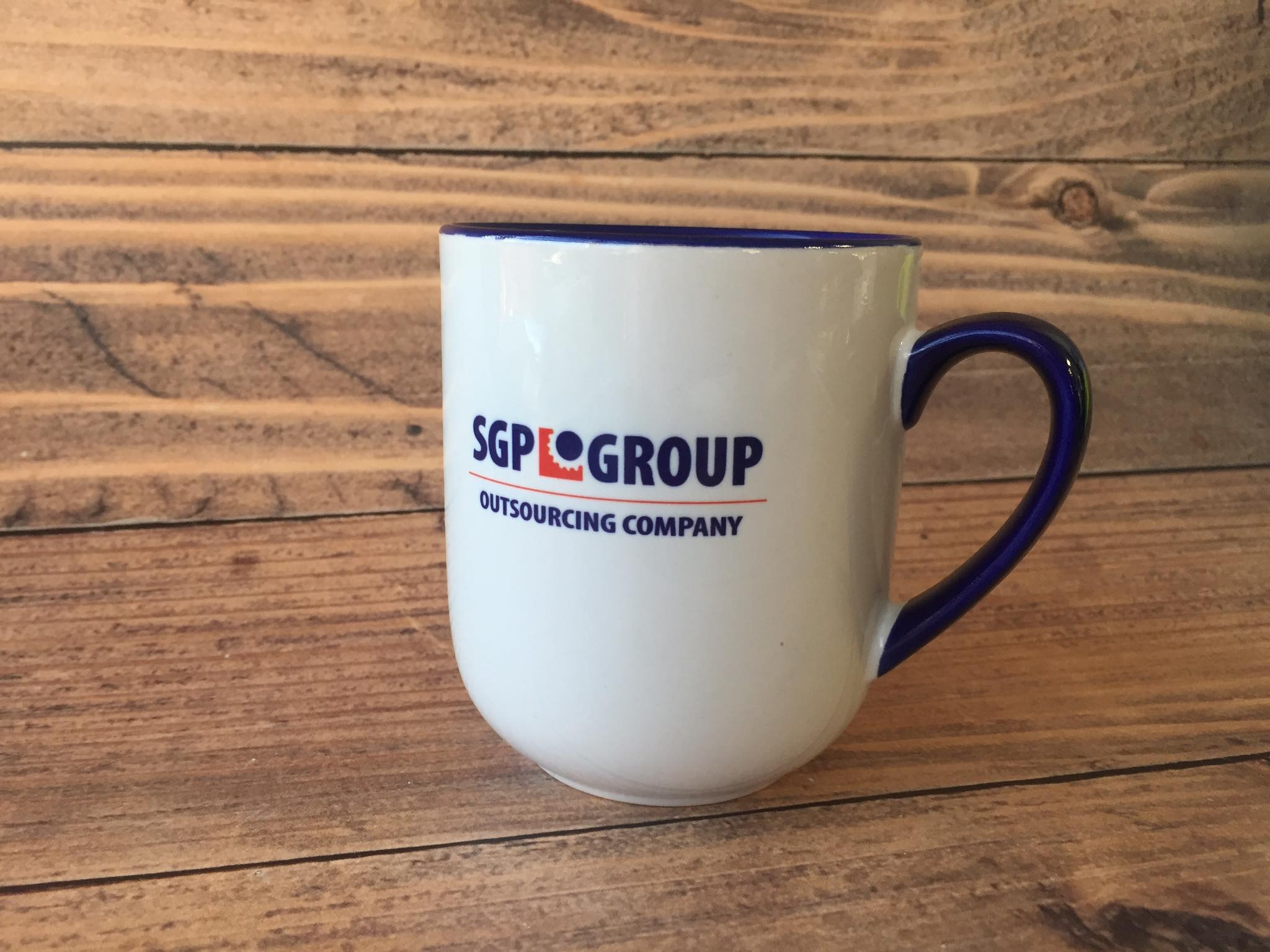 Biały kubek z niebieskim obramowaniem i logo 'SGP GROUP Outsourcing Company' na tle drewnianych desek.