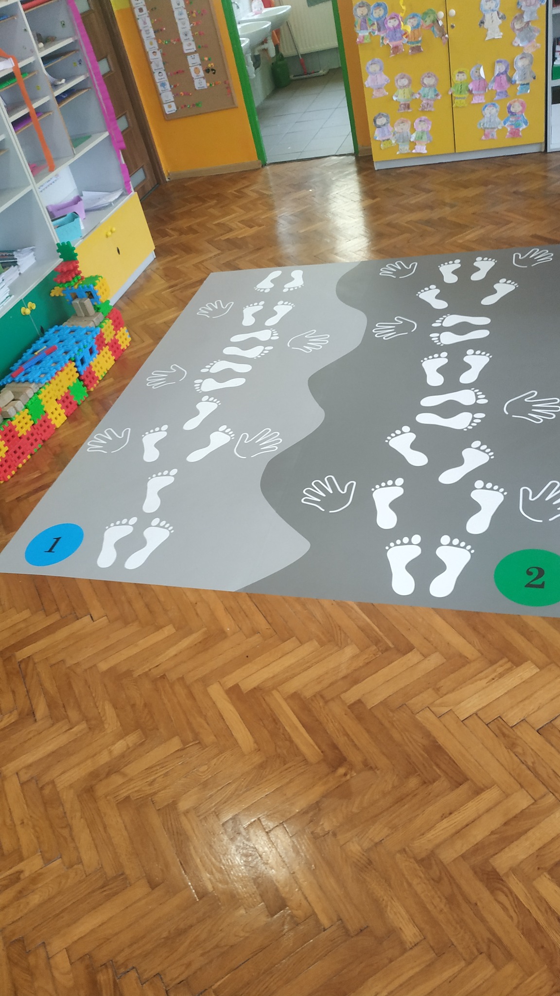 Szary dywan z białymi nadrukami stóp i dłoni, oznaczony numerami 1 i 2, leżący na parkiecie w sali przedszkolnej z kolorowymi meblami i dekoracjami.