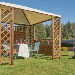 Drewniana altana z kremowym zadaszeniem, ażurowymi ścianami i stołem nakrytym do kolacji na trawniku w ogrodzie.