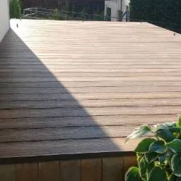 Nowoczesny taras z desek kompozytowych w Rybniku, z widocznym cieniem i roślinnością w rogu, wykonany przy jasnej ścianie budynku.