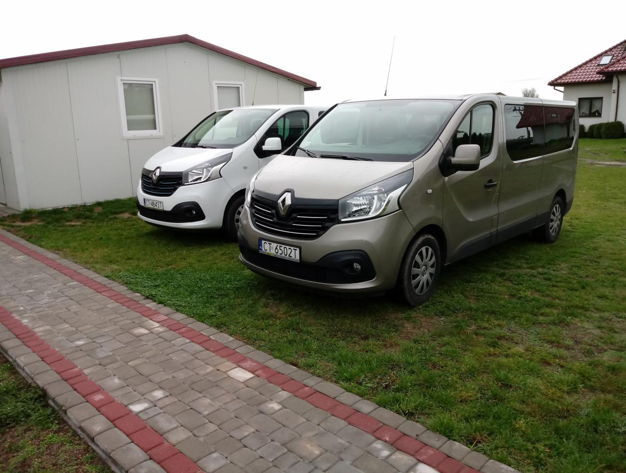 Dwa busy Renault Trafic, jeden biały, drugi beżowy, zaparkowane na trawie przed budynkiem biurowym z kostką brukową.