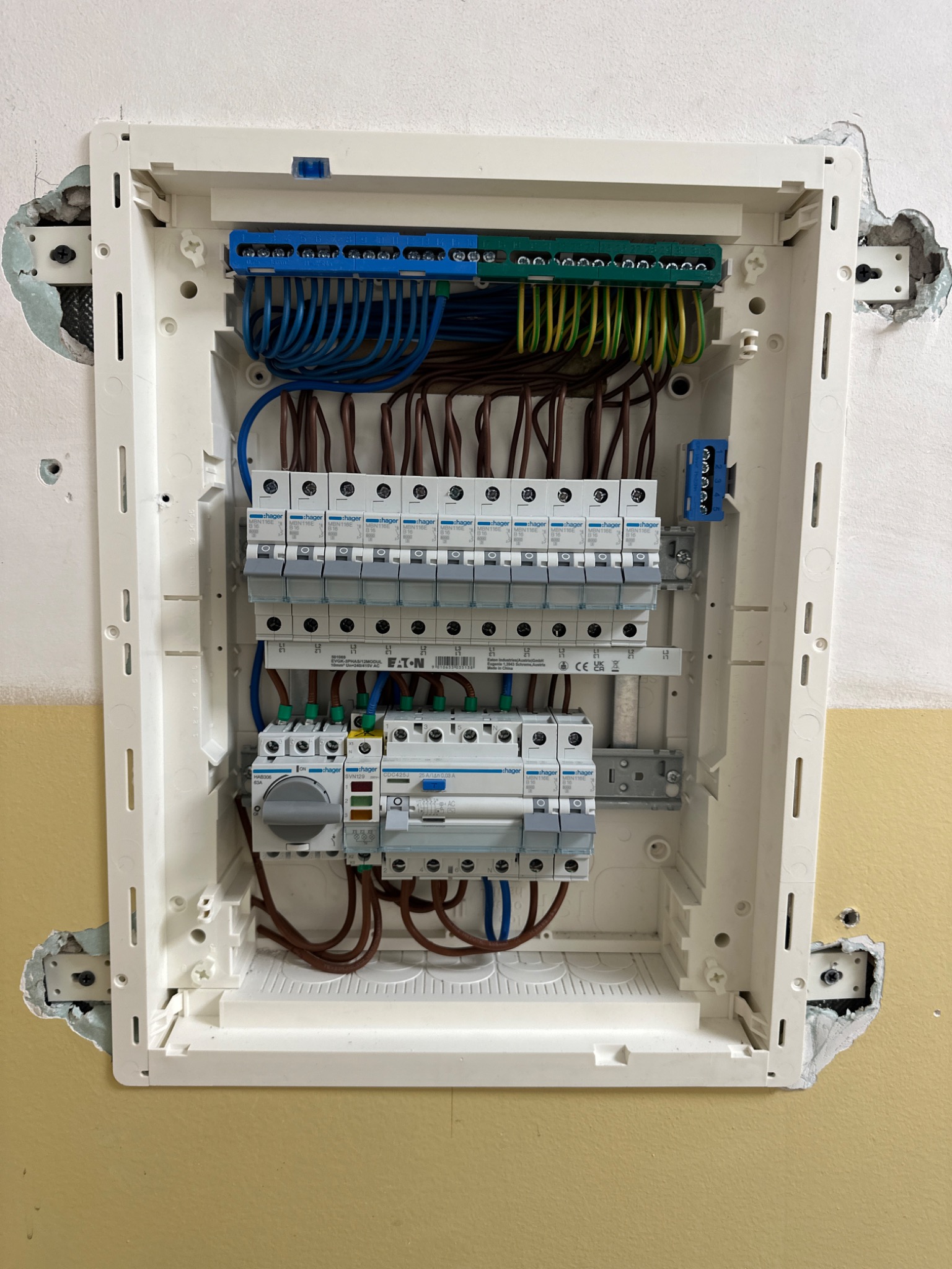 Rozdzielnica elektryczna Hager zamontowana w ścianie, widoczne bezpieczniki, przewody w kolorach niebieskim, brązowym, zielono-żółtym. Ślady po montażu na ścianie.