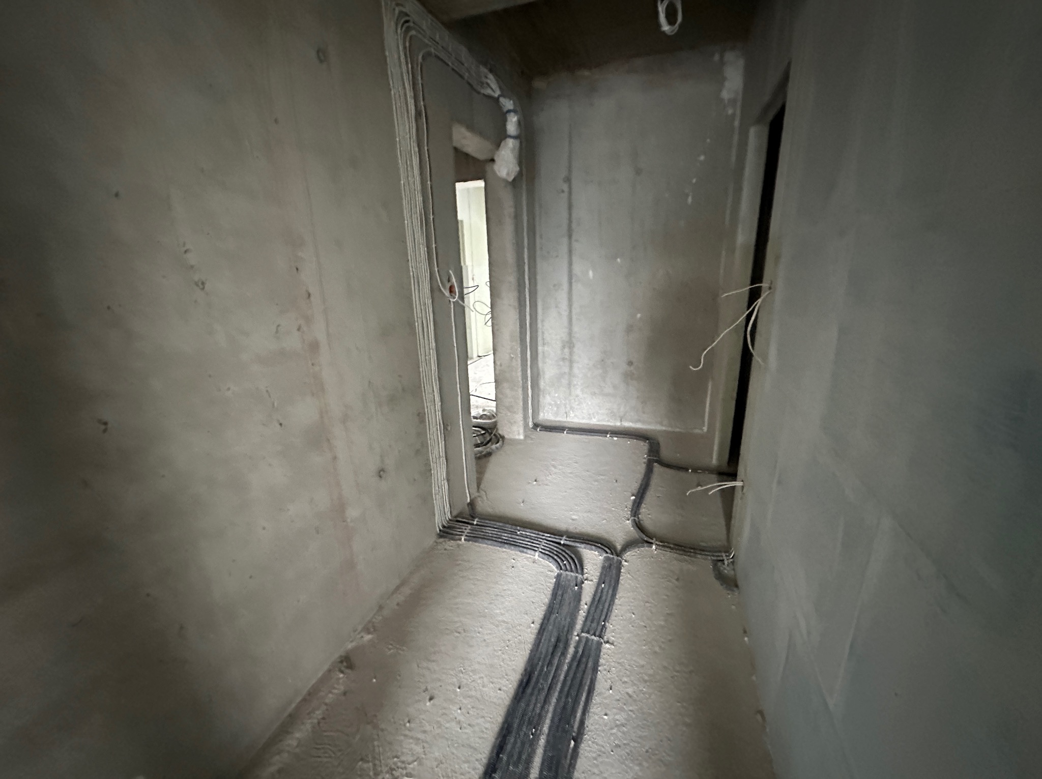 Surowy korytarz w trakcie prac wykończeniowych z widocznymi przewodami elektrycznymi ułożonymi wzdłuż ścian i podłogi. Widoczne kable w peszlach.