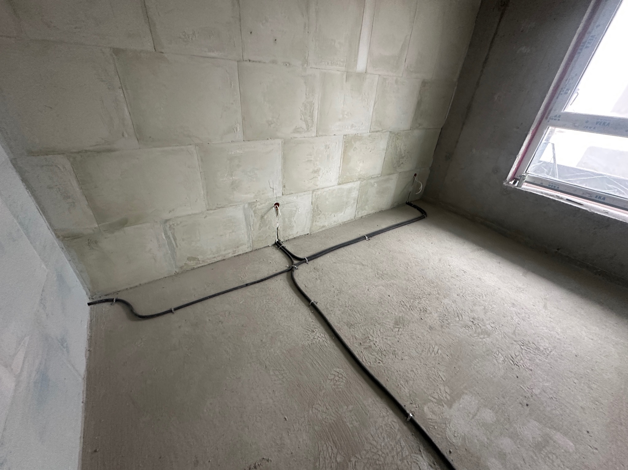 Szara ściana z widocznymi przewodami elektrycznymi, zamocowanymi do ściany. Przewody prowadzone po podłodze w surowym stanie, przed dalszymi pracami wykończeniowymi.