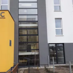 Fasada budynku z dużymi oknami w pionowym pasie, widok z zewnątrz, po lewej żółta ściana z napisem.