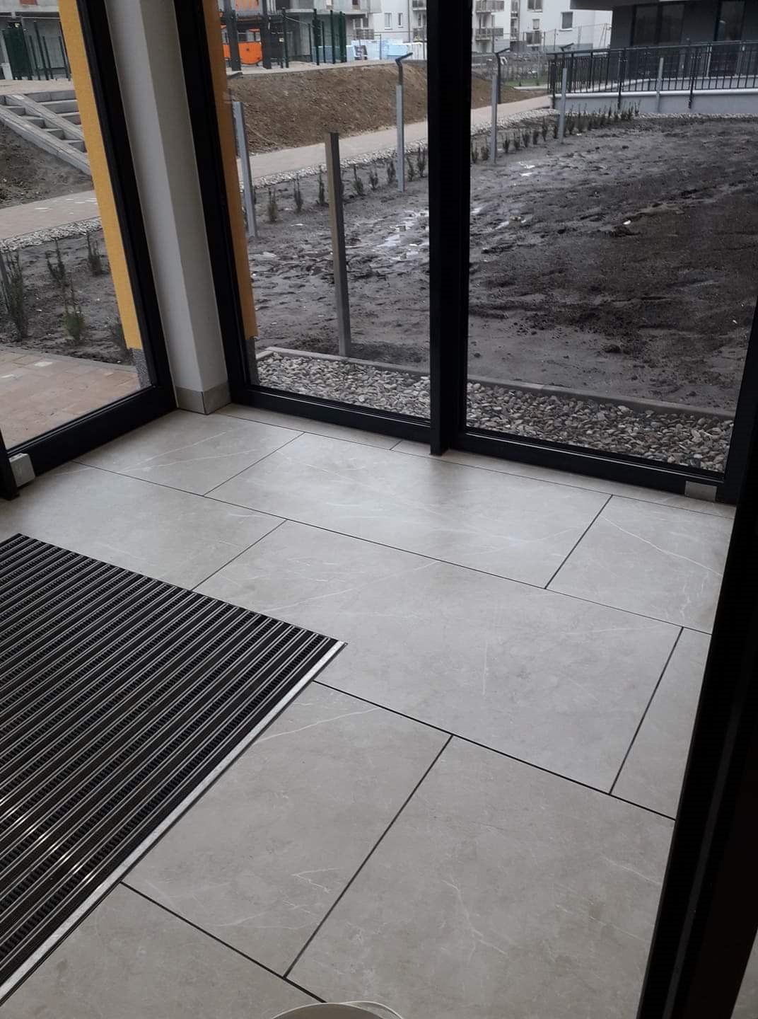 Wnętrze wejścia do budynku z szarą wycieraczką i jasnymi płytkami podłogowymi; widok na zewnątrz przez szklane drzwi ukazuje błotniste podłoże i nowo posadzone drzewka.