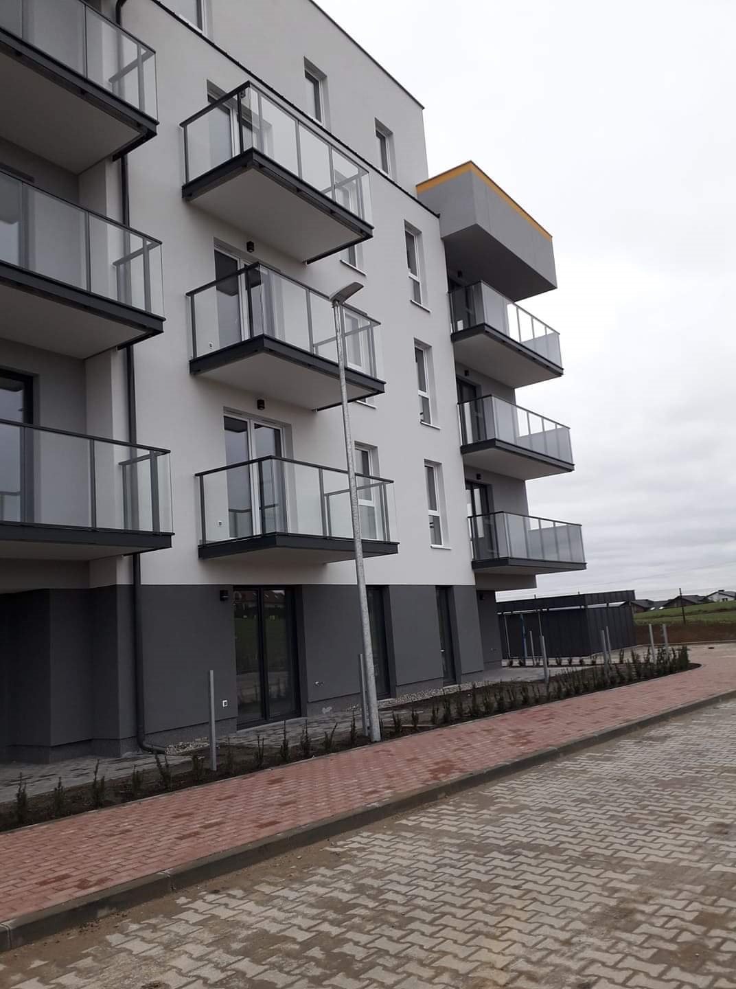Nowoczesny budynek mieszkalny z balkonami i chodnikiem z kostki brukowej, widok z poziomu chodnika w pochmurny dzień.