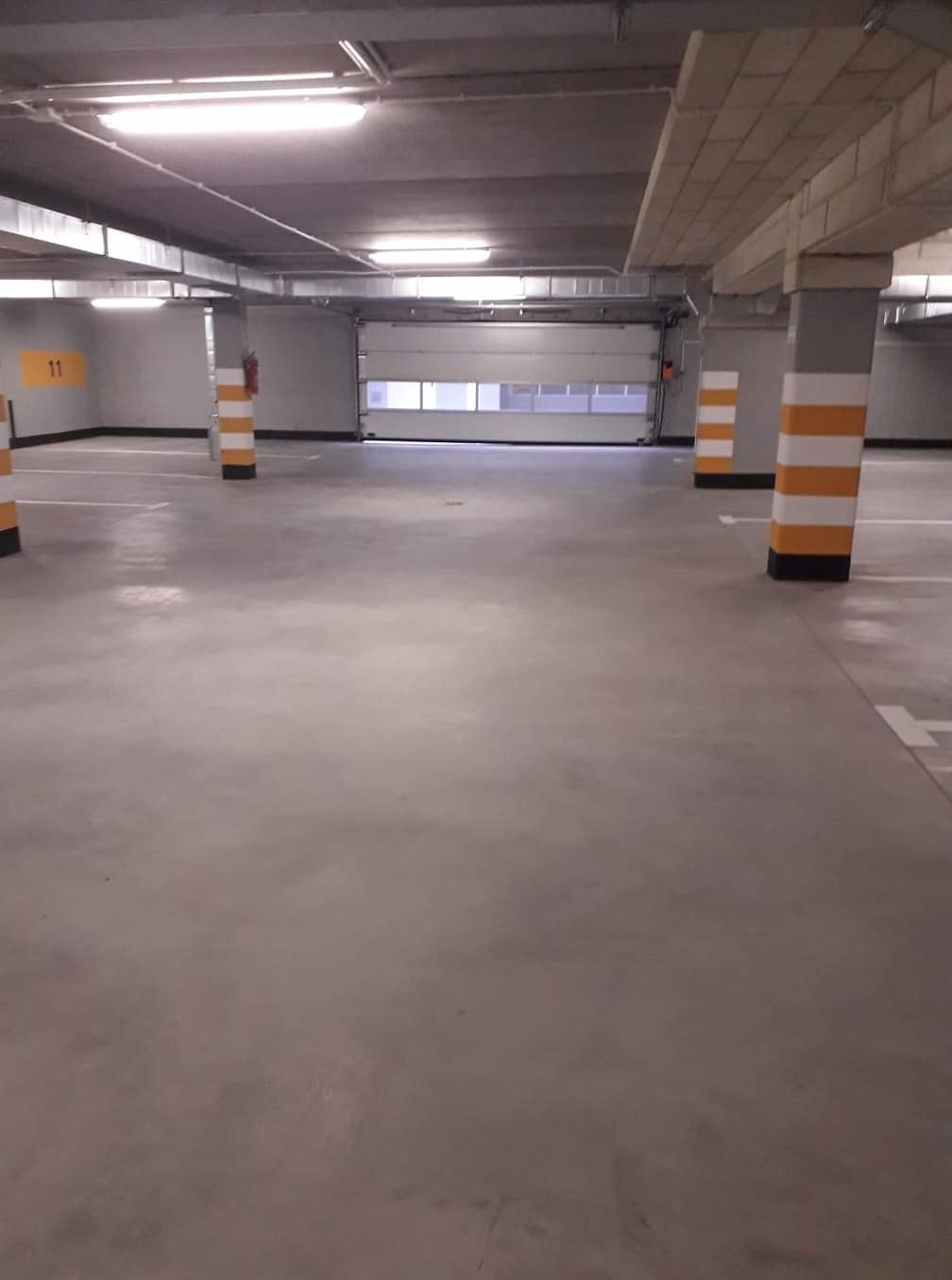 Pusty parking podziemny z betonową podłogą, oznaczonymi miejscami postojowymi i słupami pomalowanymi w żółto-czarne pasy, oświetlony lampami sufitowymi.