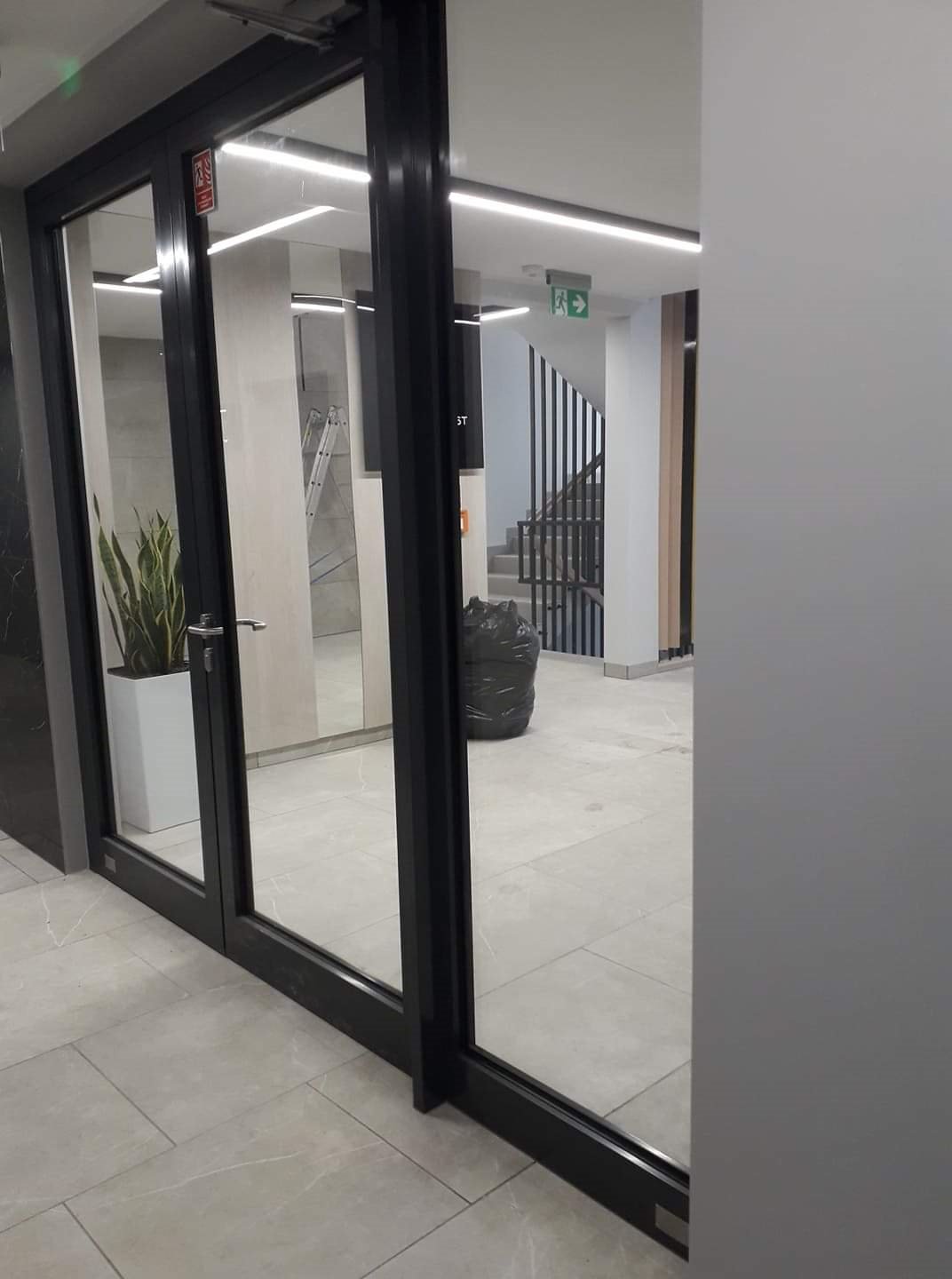 Szklane drzwi z czarną ramą w nowoczesnym budynku biurowym, widoczna roślina w donicy, schody i worek na śmieci w tle.