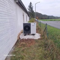 Dobry Klimat - Zewnętrzna jednostka pompy ciepła Viessmann, zamontowana na białym żwirze przy ścianie budynku z białym sidingiem, widok z boku na tle jesiennego krajobrazu.