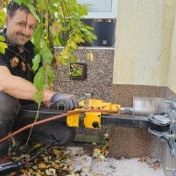 Dobry Klimat - Mężczyzna w kucki, w szarych rękawicach, wierci żółtą wiertarką rdzeniową otwór w elewacji budynku, widoczne liście drzewa w tle.