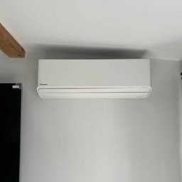 Dobry Klimat - Biała jednostka klimatyzacji Panasonic zawieszona na szarej ścianie w jasnym wnętrzu, widoczny fragment drewnianej belki stropowej i czarny telewizor z lewej strony.