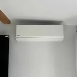 Biała jednostka klimatyzacji Panasonic zawieszona na szarej ścianie w jasnym wnętrzu, widoczny fragment drewnianej belki stropowej i czarny telewizor z lewej strony.