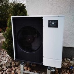 Dobry Klimat - Zewnętrzna jednostka pompy ciepła Mitsubishi Ecodan na metalowych wspornikach, otoczona kamieniami i zielenią, przy białej ścianie budynku.