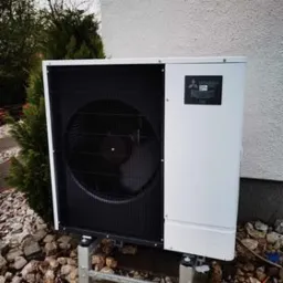 Zewnętrzna jednostka pompy ciepła Mitsubishi Ecodan na metalowych wspornikach, otoczona kamieniami i zielenią, przy białej ścianie budynku.