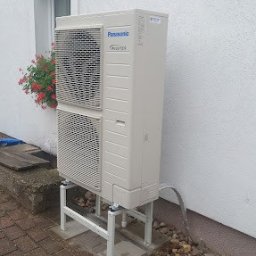 Dobry Klimat - Zewnętrzna jednostka pompy ciepła Panasonic Inverter zamontowana na metalowej konstrukcji na ceglanym chodniku, obok ściany budynku z oknem i kwiatami.
