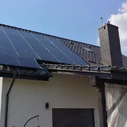 Panele fotowoltaiczne na dachu domu jednorodzinnego z ciemną dachówką, widoczne anteny satelitarne i drabina oparta o ścianę.