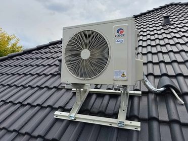 Jednostka zewnętrzna klimatyzacji Gree zamontowana na wspornikach na skośnym dachu pokrytym czarną dachówką ceramiczną, widoczne logo i oznaczenia producenta.