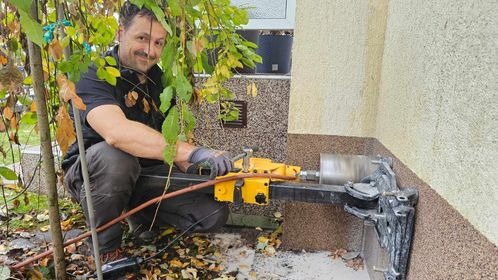 Mężczyzna w kucki, w szarych rękawicach, wierci żółtą wiertarką rdzeniową otwór w elewacji budynku, widoczne liście drzewa w tle.