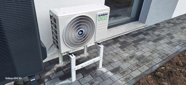Zewnętrzna jednostka pompy ciepła Kaisai Eco, zamontowana na białym stelażu przy ścianie budynku i szarej kostce brukowej. Widoczny czarny element drugiej jednostki. Świeżo ułożona kostka brukowa.