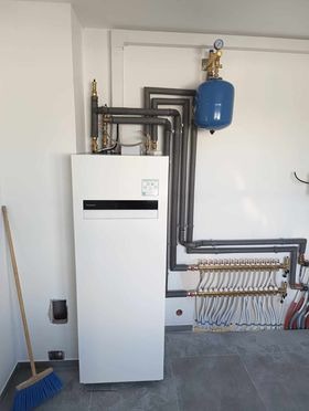 Biała pompa ciepła typu monoblok, z widocznymi rurami instalacji grzewczej, zbiornikiem wyrównawczym i rozdzielaczem podłogówki w nowym, surowym pomieszczeniu. Z boku oparta o ścianę niebieska...